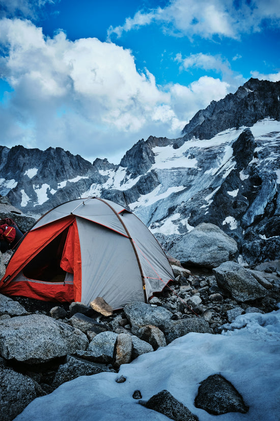 A grey and orange tent on a rocky slope below snow-capped mountains
